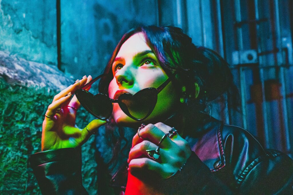 Close-up of woman with heart-shaped sunglasses and rings under colorful lights, exuding a modern, edgy vibe.