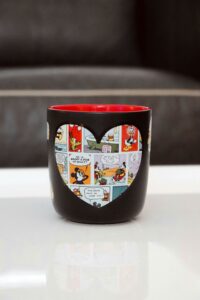 Black mug with a heart-shaped cutout revealing a colorful cartoon comic design, perfect for Valentine's Day.