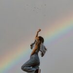 A woman in denim jeans standing under a vivid rainbow with birds in a clear sky.
